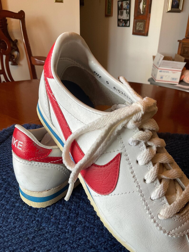 May include: White leather Nike trainers with a red swoosh and blue trim. The shoes have white laces and the text "SIZE 5 81030" is visible on the inside of the shoe.