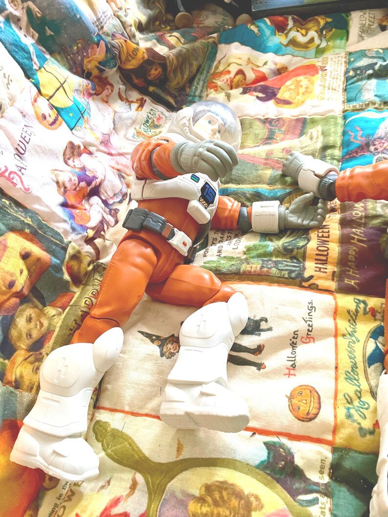 May include: An orange astronaut action figure with a clear helmet, white gloves, and boots. The figure is lying on a colorful Halloween-themed patterned fabric. The figure is holding a white object.