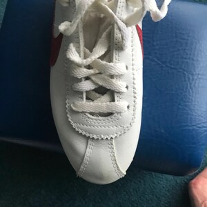 May include: White leather trainers with red accents and white laces. The shoes are tied and the laces are crossed.