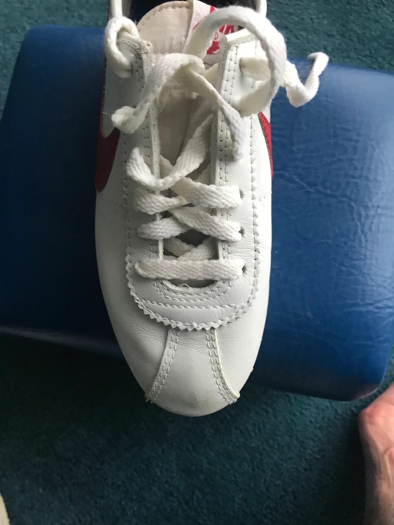 May include: White leather trainers with red accents and white laces. The shoes are tied and the laces are crossed.
