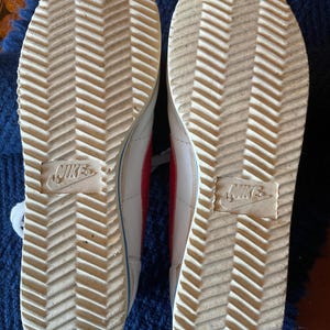 May include: White leather trainers with red accents and a herringbone pattern on the soles. The soles are marked with the Nike logo.