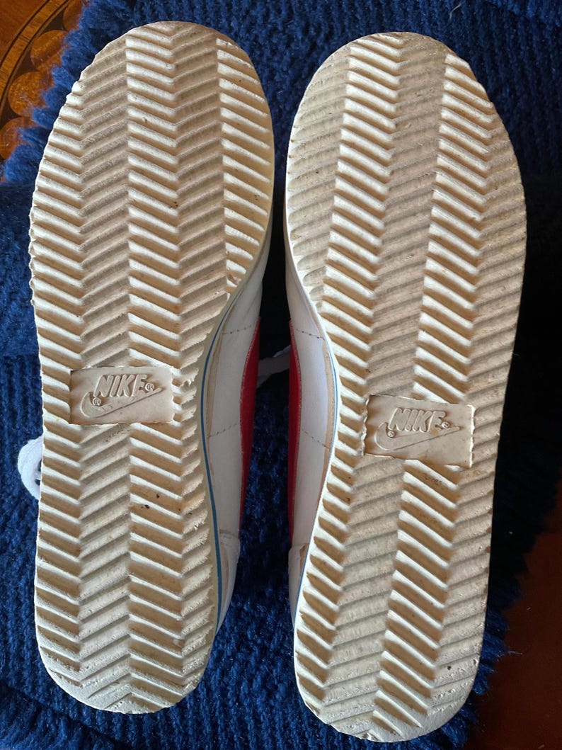 May include: White leather trainers with red accents and a herringbone pattern on the soles. The soles are marked with the Nike logo.