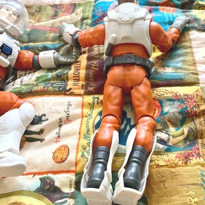 May include: Two astronaut action figures in orange suits with white accents and helmets. One figure is lying down, and the other is facing away. The figures are on a colorful Halloween-themed quilt.