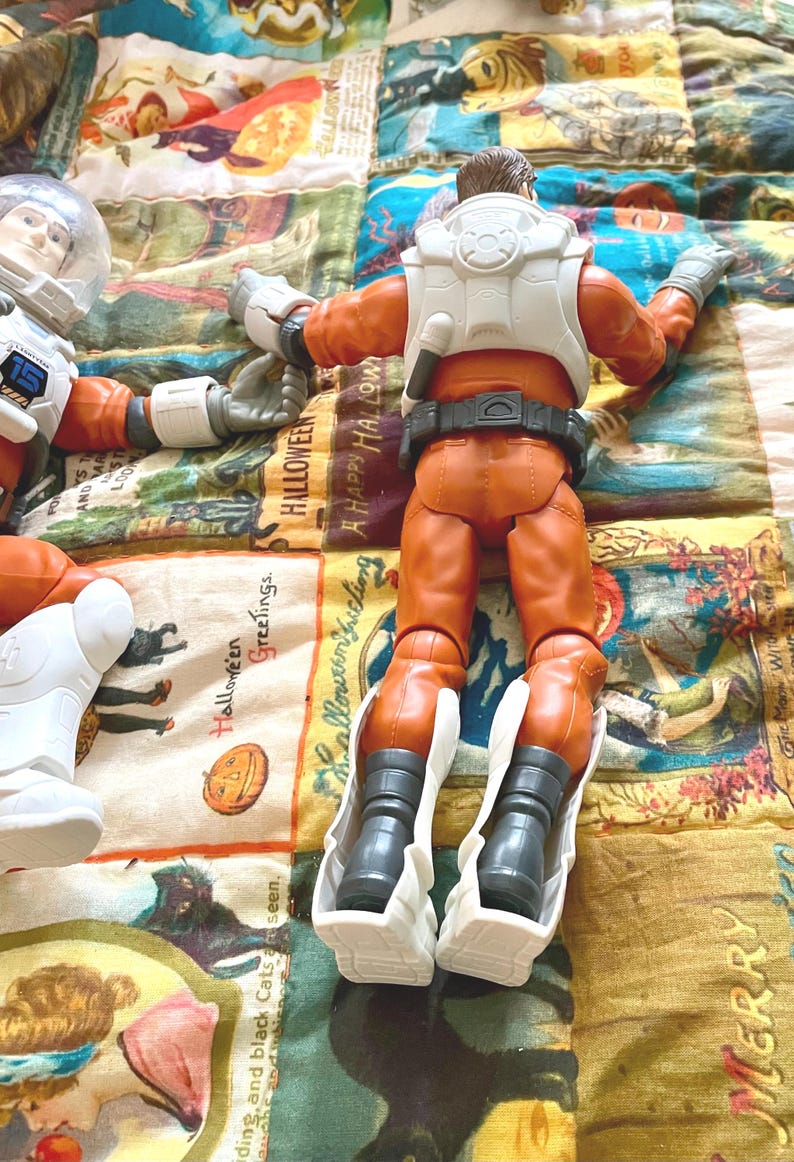 May include: Two astronaut action figures in orange suits with white accents and helmets. One figure is lying down, and the other is facing away. The figures are on a colorful Halloween-themed quilt.