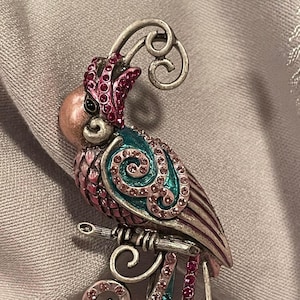 May include: A pink and teal jeweled parrot brooch with a silver filigree design. The parrot is perched on a branch with a silver swirl design.