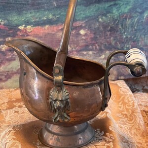 May include: A copper-toned antique sugar scuttle with a handle and a decorative lion's head. The scuttle has a small pedestal base and a swing handle. The handle on the side has a white and blue ceramic knob.