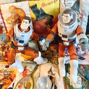 May include: Two astronaut action figures in orange and white space suits, one with a clear helmet. The figures are on a Halloween-themed fabric with pumpkins, witches, and black cats. A clear helmet and a mirror are also visible.