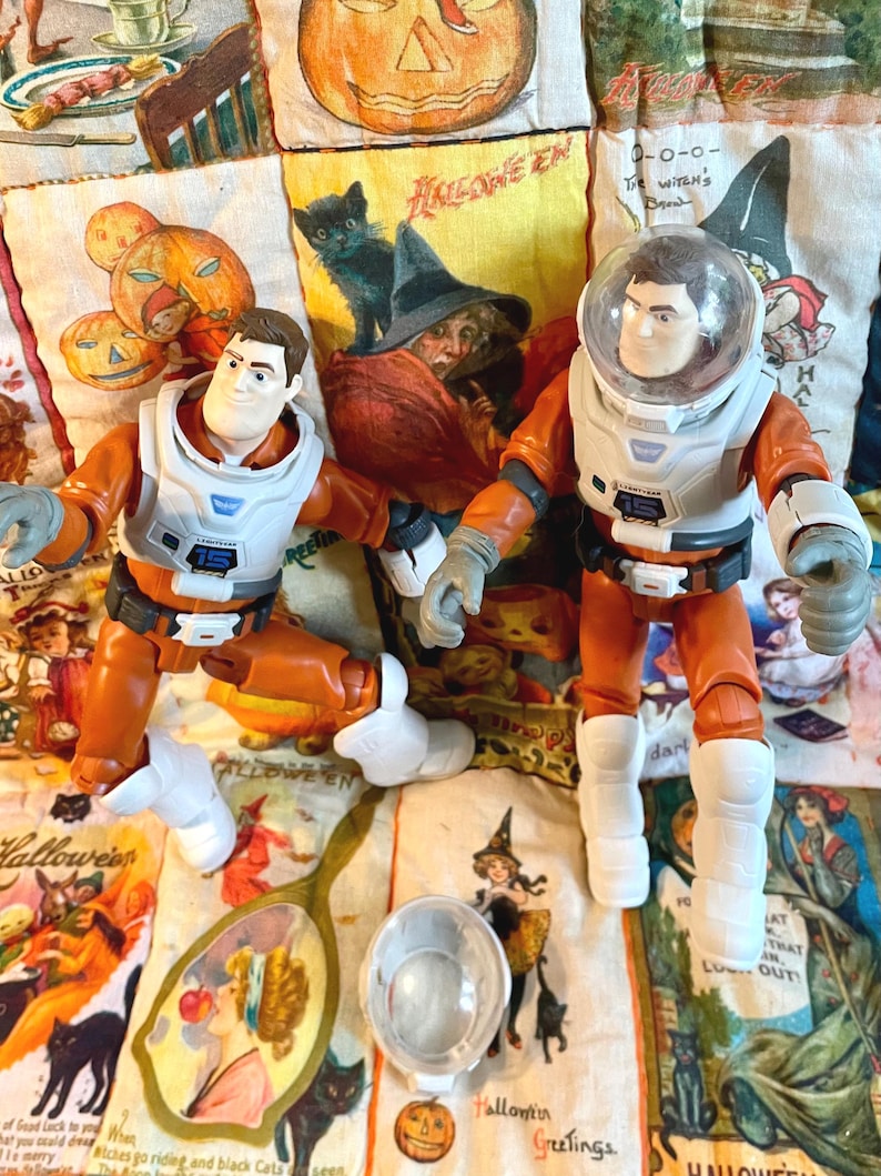 May include: Two astronaut action figures in orange and white space suits, one with a clear helmet. The figures are on a Halloween-themed fabric with pumpkins, witches, and black cats. A clear helmet and a mirror are also visible.