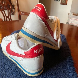 May include: A pair of white leather trainers with red and blue accents. The trainers have the word "NIKE" written in red on the back of each shoe.