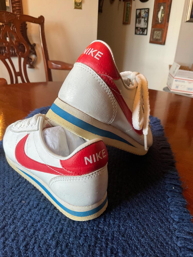 May include: A pair of white leather trainers with red and blue accents. The trainers have the word "NIKE" written in red on the back of each shoe.
