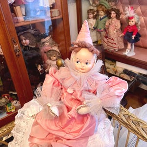 May include: A vintage doll dressed in a pink satin clown costume with a ruffled collar and matching pointed hat. The doll is seated on a white lace-covered surface, with other antique dolls visible in the background.
