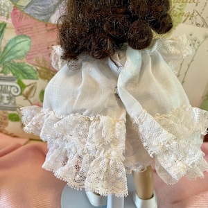 May include: A vintage doll with brown curly hair and a white dress with lace trim. The doll is standing on a white stand with a blue base. The dress has a high neckline and a ruffled skirt.