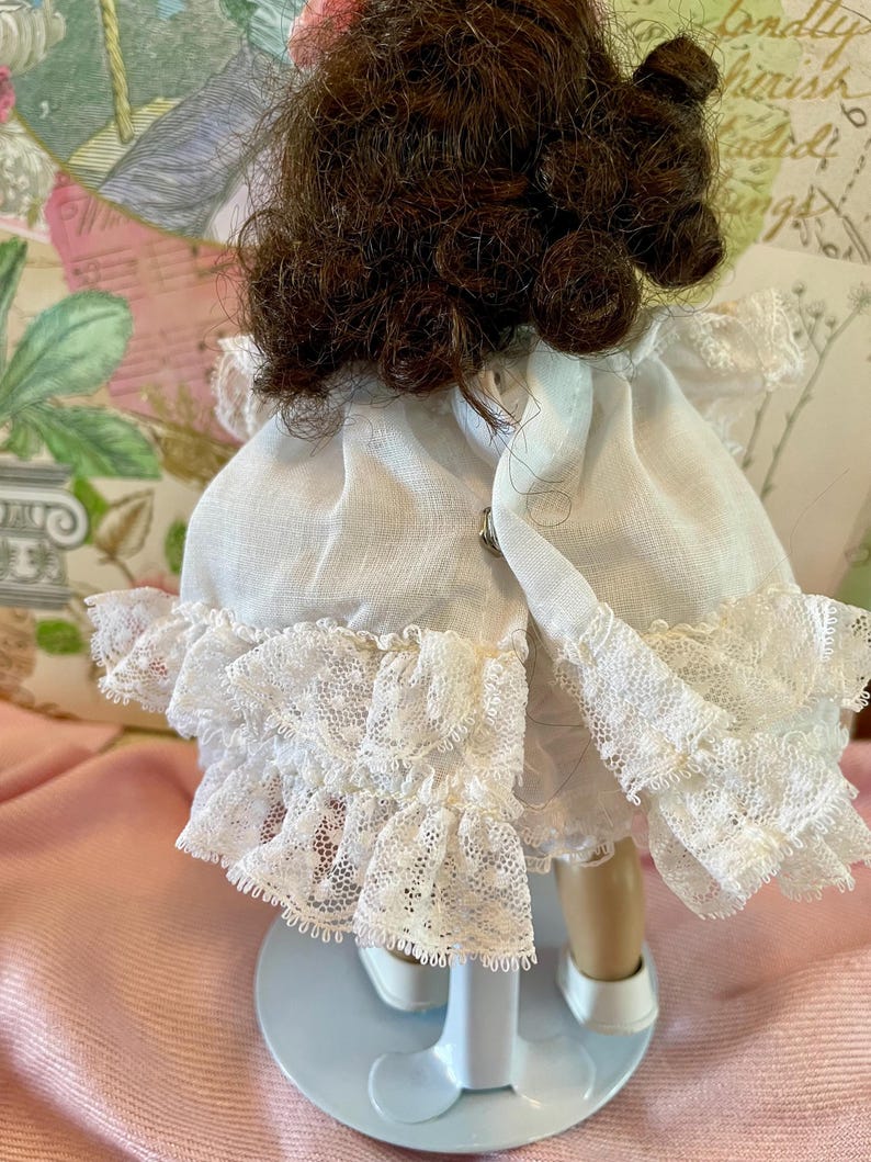 May include: A vintage doll with brown curly hair and a white dress with lace trim. The doll is standing on a white stand with a blue base. The dress has a high neckline and a ruffled skirt.