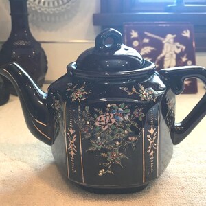 May include: A black ceramic teapot with a lid and handle. The teapot is decorated with floral designs in pink, blue, and gold. Gold accents are on the sides. The teapot is sitting on a white surface.