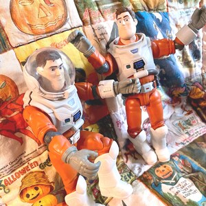 May include: Two Buzz Lightyear action figures in orange and white space suits. One figure is seated with a clear helmet, the other is standing with arms raised. Both figures have gray gloves and white boots. The figures are on a Halloween-themed quilt.