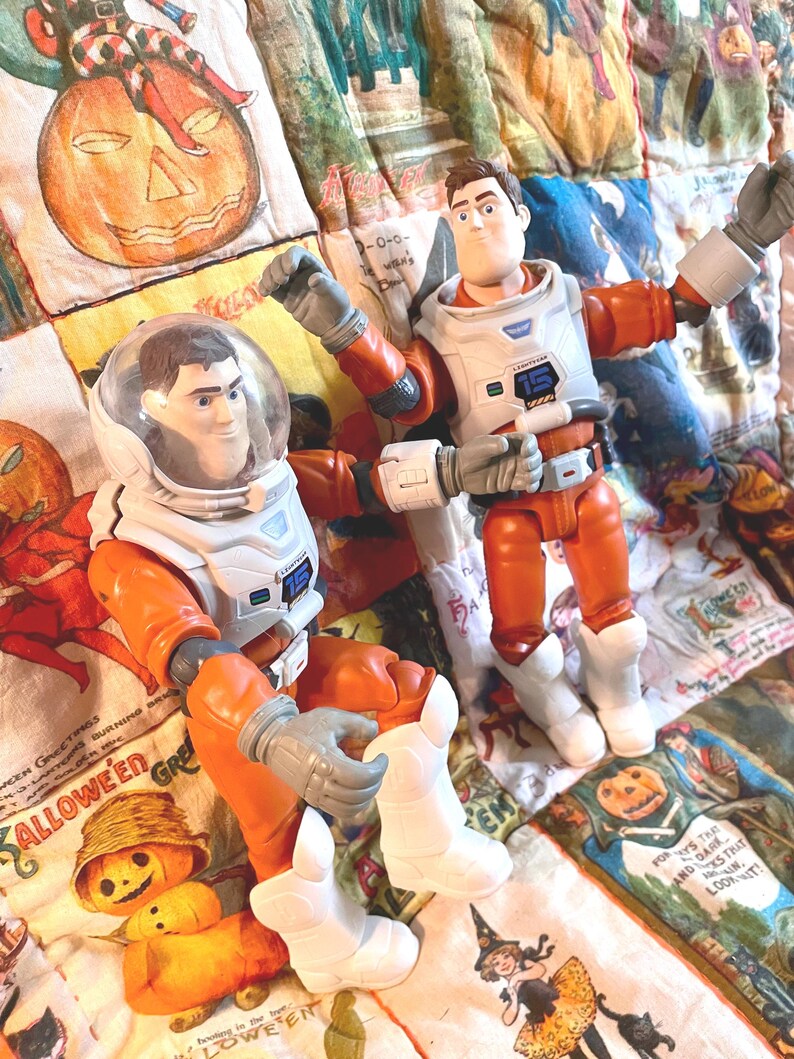 May include: Two Buzz Lightyear action figures in orange and white space suits. One figure is seated with a clear helmet, the other is standing with arms raised. Both figures have gray gloves and white boots. The figures are on a Halloween-themed quilt.