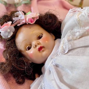 May include: A vintage doll with brown hair and a floral headband featuring pink and white roses. The doll has rosy cheeks, red lips, and is dressed in a white dress with lace trim. The doll is lying on a pink surface.