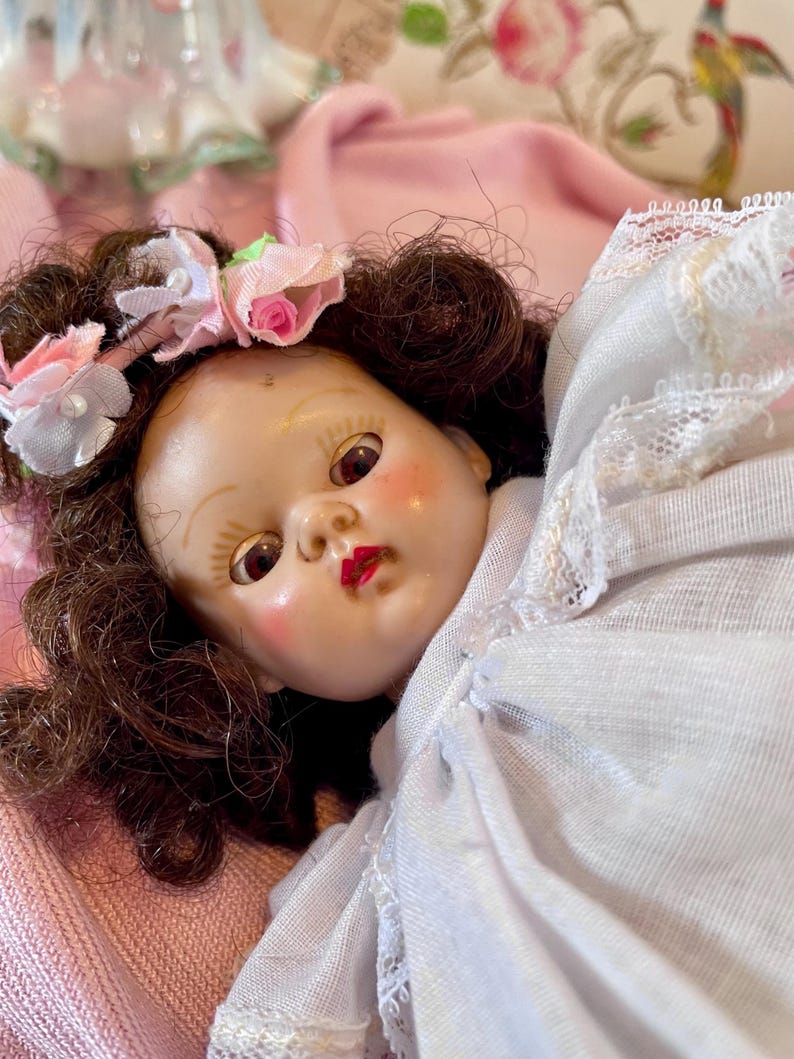 May include: A vintage doll with brown hair and a floral headband featuring pink and white roses. The doll has rosy cheeks, red lips, and is dressed in a white dress with lace trim. The doll is lying on a pink surface.