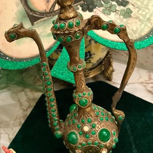 May include: A gold-colored metal teapot with a green jeweled design. The teapot has a round body and a long spout. The teapot is decorated with green gemstones.
