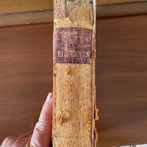 May include: Aged book spine with the title "Euclid's Elements" in gold lettering on a maroon label. The book's cover is worn and tattered, showing signs of age and use. The spine is a light brown color.
