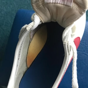 May include: Close-up of the inside of a white trainer with a red swoosh and a white tongue tag. The tongue tag has the text "THROUGHOUT THE WORLD. MPION ATHLETES NIKE AND THE SWOOSH NAME AND STRIPE ARE TRADEMARKS AND YOUR" printed on it.