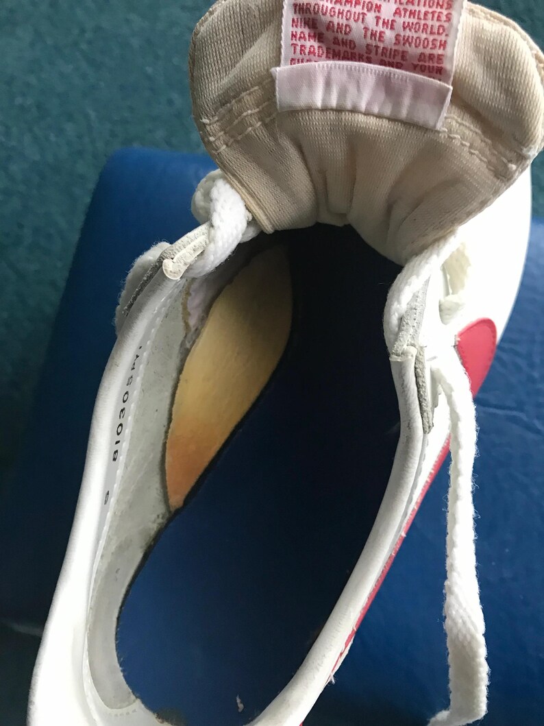 May include: Close-up of the inside of a white trainer with a red swoosh and a white tongue tag. The tongue tag has the text "THROUGHOUT THE WORLD. MPION ATHLETES NIKE AND THE SWOOSH NAME AND STRIPE ARE TRADEMARKS AND YOUR" printed on it.