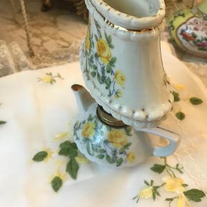 May include: A white porcelain teapot with a yellow and green floral design. The teapot has a lid that resembles a lampshade with a gold trim. The teapot is sitting on a white tablecloth with yellow floral embroidery.
