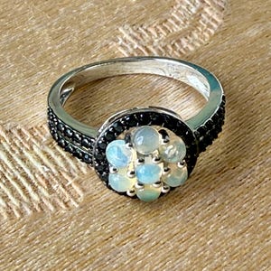 May include: A silver ring with a cluster of light blue and white gemstones at the center. The ring has a black band around the gemstones and black stones on the sides. The ring is set against a textured, tan background.