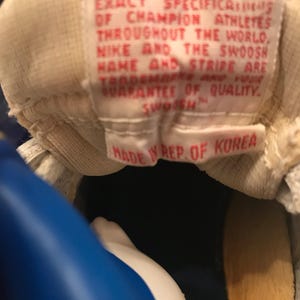 May include: Close-up of a white label with red text that reads "NIKE SPORT SHOES ARE MANUFACTURED TO THE EXACT SPECIFICATIONS OF CHAMPION ATHLETES THROUGHOUT THE WORLD, NIKE AND THE SWOOSH NAME AND STRIPE ARE TRADEMARKS AND YOUR GUARANTEE OF QUALITY. SWOOSH. MADE IN REP. OF KOREA."