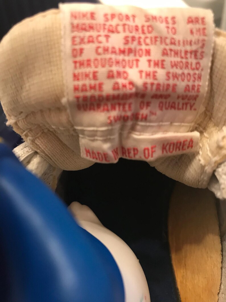 May include: Close-up of a white label with red text that reads "NIKE SPORT SHOES ARE MANUFACTURED TO THE EXACT SPECIFICATIONS OF CHAMPION ATHLETES THROUGHOUT THE WORLD, NIKE AND THE SWOOSH NAME AND STRIPE ARE TRADEMARKS AND YOUR GUARANTEE OF QUALITY. SWOOSH. MADE IN REP. OF KOREA."