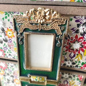 May include: A small, rectangular photo frame with a green enamel finish and gold-colored trim. The frame has a decorative floral and scroll design at the top and around the edges. The background features floral patterned paper.
