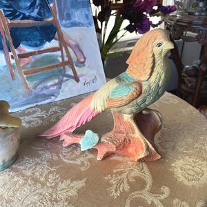 May include: A ceramic bird figurine perched on a textured branch. The bird features a soft color palette of peach, light blue, and cream, with detailed feather textures. The branch is colored in shades of peach and brown with a single light blue leaf.
