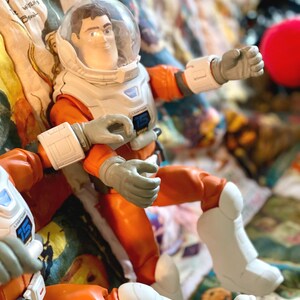 May include: A Buzz Lightyear action figure in an orange and white spacesuit with a clear helmet. The figure has gray gloves and white boots. The figure is posed on a colorful patterned surface.