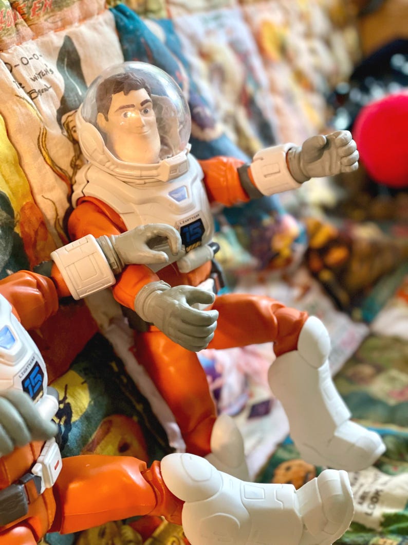 May include: A Buzz Lightyear action figure in an orange and white spacesuit with a clear helmet. The figure has gray gloves and white boots. The figure is posed on a colorful patterned surface.