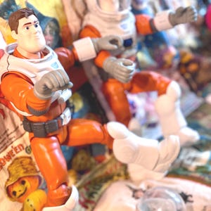 May include: Two action figures of an astronaut in an orange and white spacesuit. The figures have gray gloves and white boots. One figure is in a seated position, and the other is standing. The background is a patterned fabric with Halloween-themed images.