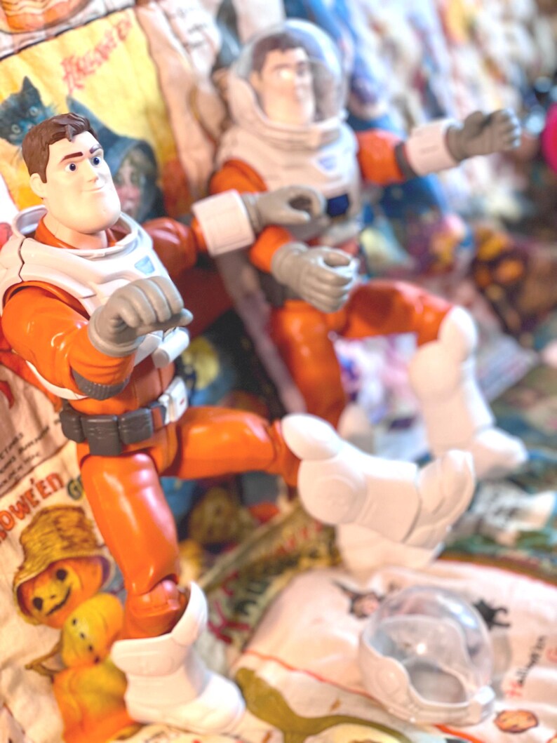 May include: Two action figures of an astronaut in an orange and white spacesuit. The figures have gray gloves and white boots. One figure is in a seated position, and the other is standing. The background is a patterned fabric with Halloween-themed images.