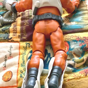 May include: An orange action figure wearing a white space suit and boots. The figure is lying on a colorful Halloween-themed patterned fabric. The figure has a black belt and a white backpack.