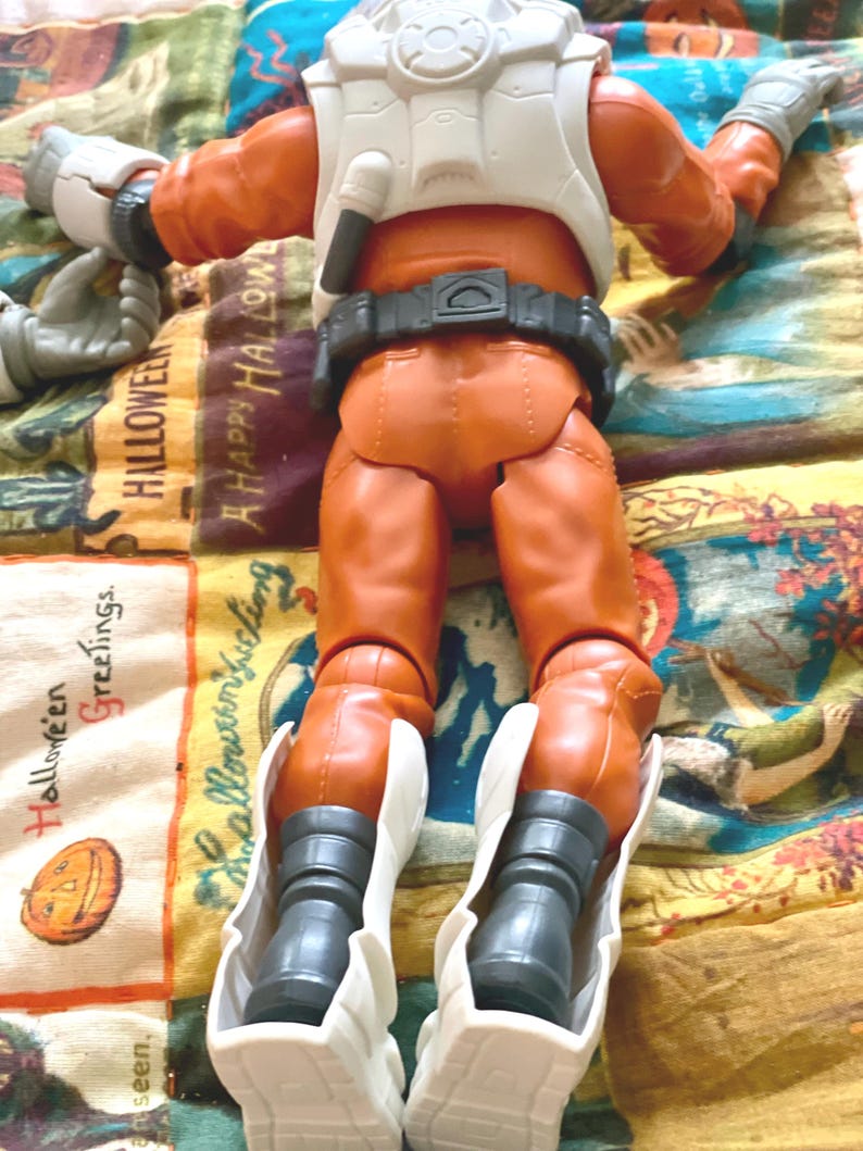 May include: An orange action figure wearing a white space suit and boots. The figure is lying on a colorful Halloween-themed patterned fabric. The figure has a black belt and a white backpack.