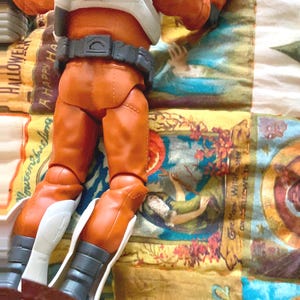 May include: An action figure wearing orange pants, a white vest, and a gray belt. The figure has gray gloves and white accents on the boots. The figure is on a colorful patterned fabric.