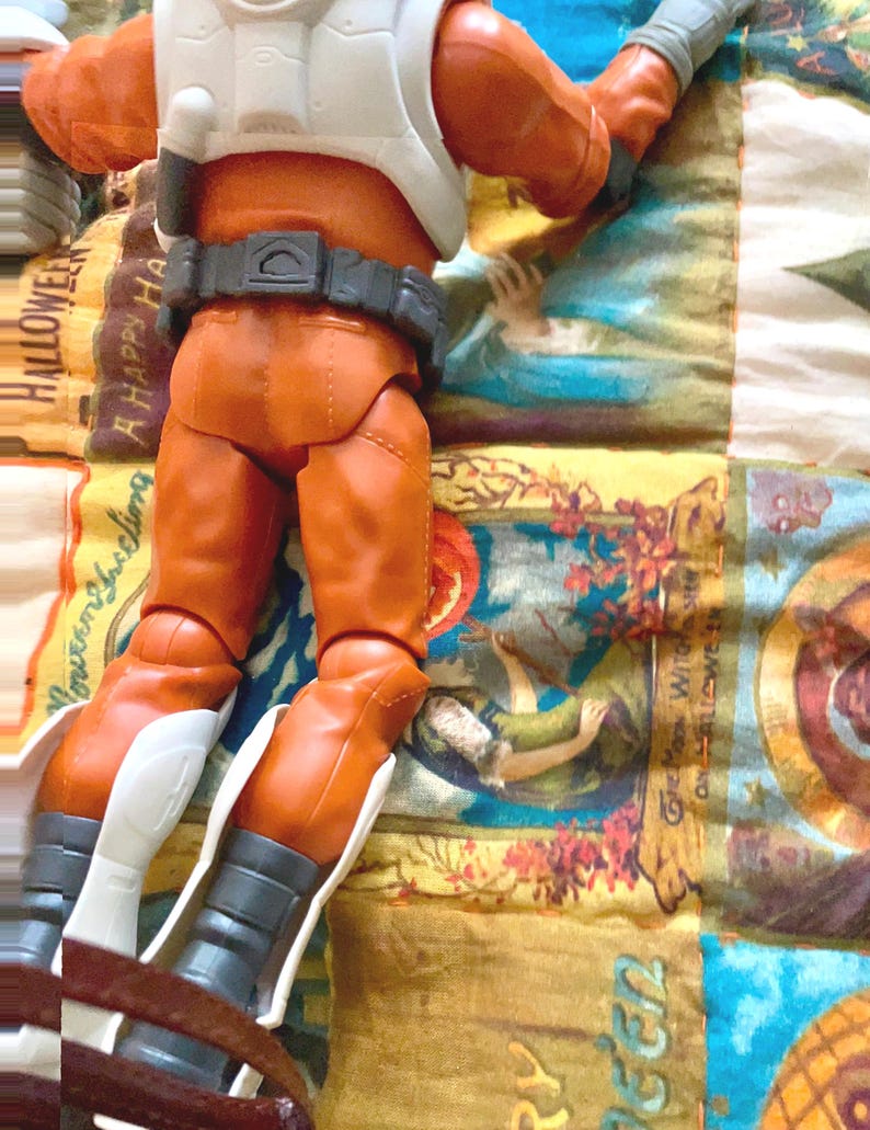 May include: An action figure wearing orange pants, a white vest, and a gray belt. The figure has gray gloves and white accents on the boots. The figure is on a colorful patterned fabric.