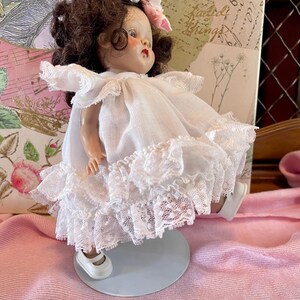 May include: A vintage doll with brown hair and pink floral accents. The doll wears a white dress with lace trim and white shoes. The doll is on a light blue stand, set against a pink background.