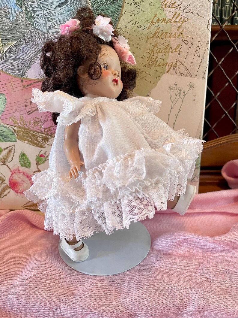 May include: A vintage doll with brown hair and pink floral accents. The doll wears a white dress with lace trim and white shoes. The doll is on a light blue stand, set against a pink background.