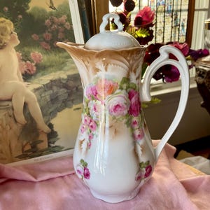May include: A white porcelain pitcher with a lid, adorned with pink and red roses and green leaves. The pitcher has a brown and tan gradient at the top and a white handle. The pitcher is a decorative item.