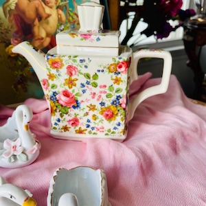 May include: A square, floral-patterned teapot with a matching lid. The teapot features a white base adorned with pink, yellow, and blue flowers. Gold trim accents the edges. Swan-shaped decorative pieces are visible in the foreground.