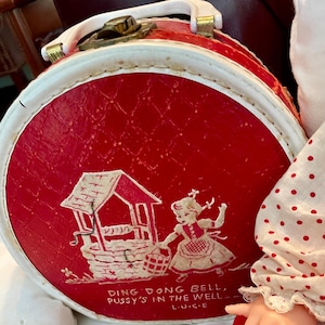 May include: A vintage red and white round suitcase with a handle and clasp. The suitcase features a graphic of a girl near a well, with the text "DING DONG BELL, PUSSY'S IN THE WELL". The suitcase has a white trim.