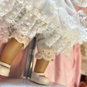 May include: A vintage doll with tan legs and white shoes. The doll is wearing white lace trimmed bloomers and a white dress with lace detailing. The doll is on a metal stand, with a pink background.