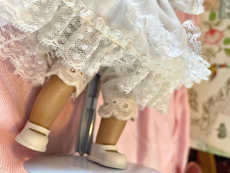 May include: A vintage doll with tan legs and white shoes. The doll is wearing white lace trimmed bloomers and a white dress with lace detailing. The doll is on a metal stand, with a pink background.