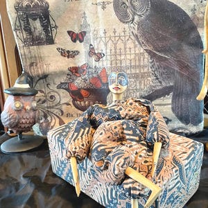 May include: A decorative doll with a painted face and wooden limbs, dressed in patterned fabric, sits on a matching box. Behind it is a canvas with an owl illustration and the text "Twisted Twigs Garden". An owl figurine with a witch hat is also visible.