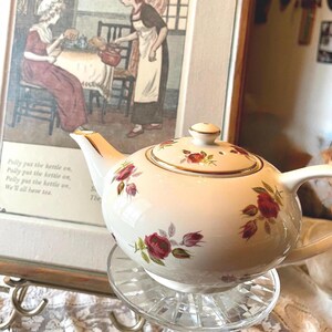 May include: A white porcelain teapot featuring a delicate red rose and green leaf pattern with gold trim on the spout, handle, and lid. The teapot has a rounded body and a matching lid with a small knob, showcasing a floral design.