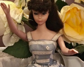 May include: A vintage Barbie doll wearing a blue and white striped dress with white lace trim. The doll has brown hair and blue eyes. The doll is posed with one arm raised and the other hand resting on a surface. The doll is surrounded by yellow roses.